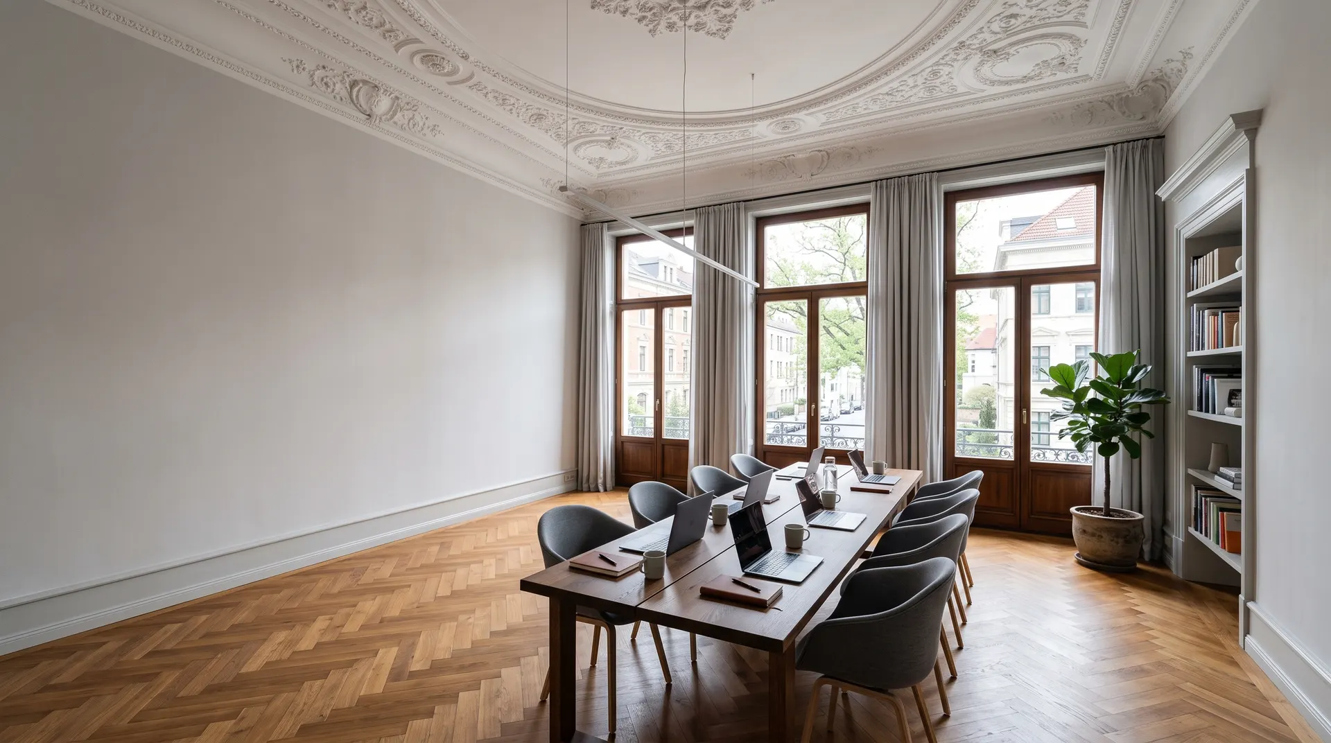 Heller, hoher Arbeitsraum in einer Villa mit Stuckdecke, Parkettboden, großen Fenstern und einem langen Holztisch mit grauen Stühlen.